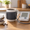 Person misst den Blutdruck neben einer Tasse Kaffee und einem Blutdruckmessgerät auf dem Tisch